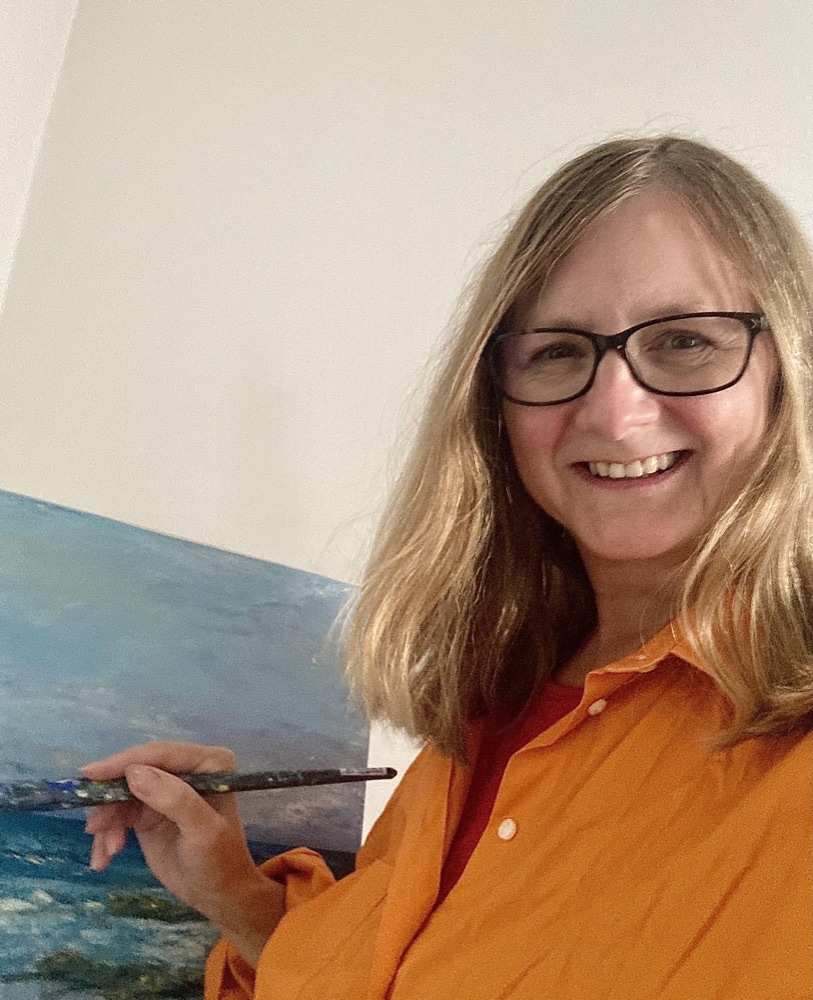 Emma Lightowlers in her studio, holding a brush beside one of her seascapes